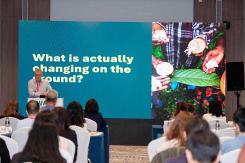 Peter Feilberg presents to an audience at the WAFI conference, beside a slide reading &ldquo;What is actually changing on the ground?&rdquo; with images of cocoa pods and farmers&rsquo; hands.